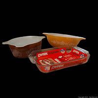 Set of four Pyrex glass baking and mixing pieces, including an oblong clear dish with packaging, a brown floral covered casserole, and two mixing bowls in brown and orange.