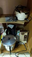 Photo showing General Electric can opener on a shelf beside Toshiba white rice cooker with cord coiled and various metal kitchen items below including a popcorn maker and Proctor Silex kettle