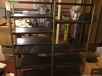 Metal shelving unit with five black metal shelves supported by light-colored metal frame, placed in a basement with various stored boxes and tools around.