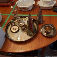 Photo showing full lot on wood table; large round silver plated tray, silver plated cone container with cap, mug, small decorative plates, salt and pepper shakers, small lidded container, and additional small dishes visible.