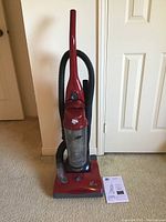 Front view of red Dirt Devil upright vacuum cleaner with clear dust compartment and attached hose, next to owner's manual on carpeted floor.