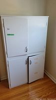 Front view of white kitchen cabinet with four doors, positioned against wall on hardwood floor. One door missing handle.