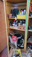 Shelving unit with black rubber boots on top shelf, assorted chemical bottles including insecticides and rat poison, fertilizers on middle shelf, white buckets and gardening accessories on bottom shelf.