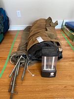 Canvas tent packed in brown duffel bag alongside tent poles and Rayovac Workhorse lantern on wooden floor.