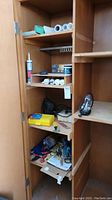 Wooden shelving cabinet with various tools, hardware, caulk tubes, stapler, paint scrapers, small tool box and other items.