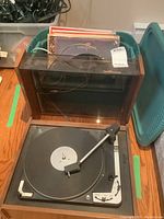 Photo showing the Dual 1010 turntable with clear hinged lid, wooden case, and a collection of vinyl records behind it.