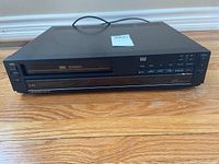 Front angle of black LXI VHS player on wooden floor showing tape slot and controls.
