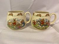 Photo showing two rounded Royal Doulton Bunnykins mugs side by side, each decorated with colorful bunny scenes and the running bunny motif around the rim.