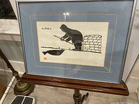 Framed print displayed on wooden stand with lamp and floor visible, showing the full artwork and matting.