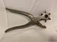 Full view of vintage metal leathercraft hole punch tool with curved handles and rotating punch wheel.