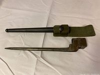 Bayonet alongside black metal scabbard with attached green fabric frog with snap button.