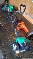 Two green weedeaters and one orange small chainsaw arranged on a wooden surface, all appear used and in need of repair or parts.