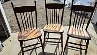 Three small wooden chairs with spindle backs and natural wood contoured seats and turned legs shown outdoors in sunlight.
