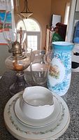 Hurricane oil lamp, dinner plates, three vases including blue floral one, and white gravy boat grouped on a surface
