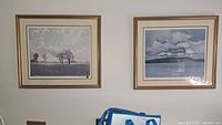 Two framed prints hanging on a wall: left print shows a tree landscape "Spring Breeze," right print a lake scene "Saw Mill".
