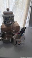 Photo showing ceramic biscotti jar, small saucepan, single wine glass, and white Canada mug with kitchen gadgets on a white counter.