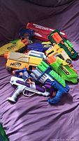 Photo of various handheld Nerf and air blasters, untested condition, arranged on a purple fabric surface