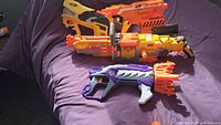 Photo showing three different Nerf blasters on purple fabric: yellow and orange Vulcan EBF-25 with belt drum, yellow and orange Ravager, purple and white N-Strike Elite blaster with orange dart holders.