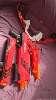 Photo showing four Nerf and Rival toy blasters including Mega Lightning Bow, Barrel Break 1X-2, Rival XVI-1200, and Rival XV-700 in red, orange, black colors on a purple surface.