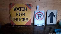 Three older traffic signs photographed on a wooden surface, including a large yellow 'WATCH FOR TRUCKS' sign, a 'No Parking' sign, and an upward arrow sign.