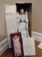 Mother doll in blue satin dress alongside baby doll in pink dress and certificate of authenticity, standing with original packaging.