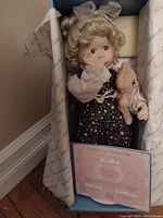 Front view of the doll in original box holding teddy bear with certificate of authenticity visible