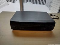 Photo of black Sharp VC-H811 VCR unit on wooden surface with visible front control panel and power cord at the back.