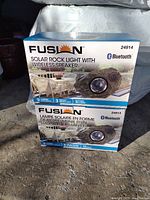 Two unopened boxes of Fusion solar rock light Bluetooth wireless speakers stacked outdoors on a concrete surface.