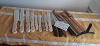 Photo of 12 cutting utensils with white handles, 3 wooden spoons, and 1 wooden ruler arranged on a table with a yellow tablecloth.