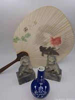 Fan with wooden handle and painted koi fish on paper surface, displayed with two carved foo dogs and a blue and white vase.