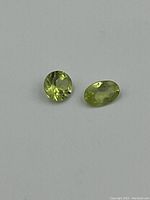 Photo of two loose faceted peridot gemstones on a white background. One is round cut, the other oval cut. Both light green color.