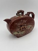 Front angled view showing the oxblood stone surface, animal finial on lid, floral carving on the front side, and spout.