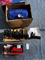 Blue air tank with pressure gauge in a worn cardboard box, new black strut with coil spring, and boxed Airtex fuel pump on a concrete surface.