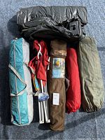 Multiple folded tents and camping chairs with cases laid on a carpet floor showing various colors and materials