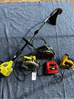 Full lot photo showing 5 power tools laid on gray fabric surface: Ryobi weed eater, two circular saws, jigsaw and sander