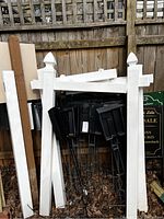 White wooden yard posts with pointed tops, black metal ground stakes, and white boards leaning against a wooden fence outside.