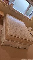 Double bed with floral patterned mattress and white bed skirt in a room with beige carpet and window in the background
