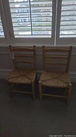Two small wooden kids chairs with woven straw seats placed side by side near window blinds.
