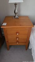 Front view of one solid wood nightstand featuring three drawers with round wooden knobs and a silver lamp on top.