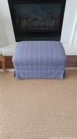 Front view of rectangular blue upholstered ottoman with white stripes on a rug, in front of a fireplace.