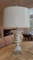 Full view of porcelain table lamp showing white drum lampshade and urn-shaped base with gold accents on a beige marble table.