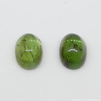 Two oval cabochon green tourmalines side by side on white background showing inclusions and color.