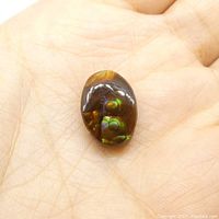 Fire agate oval cabochon gemstone resting on palm to show scale, displaying brown base with iridescent green and orange flashes