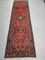 Full-length view of rug laid flat on white floor showing red field and border pattern