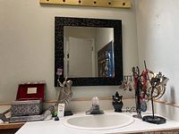 Overview of lot items on white bathroom countertop including jewelry stands, soapstone figurine, jewelry box, hand mirror, and small home decor items