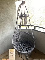 Gray macrame hanging swing chair with fringe tassels suspended from a curved metal stand on a balcony.