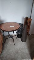 Side view of round concrete and tile table with metal frame alongside black metal umbrella stand with cut-out design, hexagonal bamboo stand, and tall amber vase corner visible.