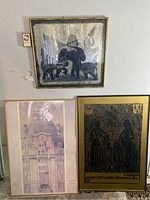 Three framed wall art pieces mounted on a wall: a dark blue monochrome print of a mythical figure with animals, a sepia/pinkish architectural drawing, and a black and gold medieval figures print.