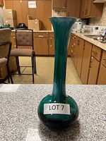 Tall bud vase with rounded bottom, deep green glossy glaze, narrow neck with flared rim, placed on countertop in kitchen setting.