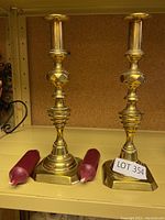 Photo showing pair of brass candlesticks set next to two red taper candles on a corkboard background.